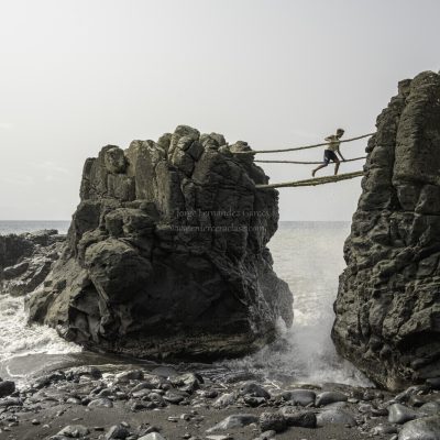 Boy crossing a precarious bridge made of logs while the waves beat on the rocks.